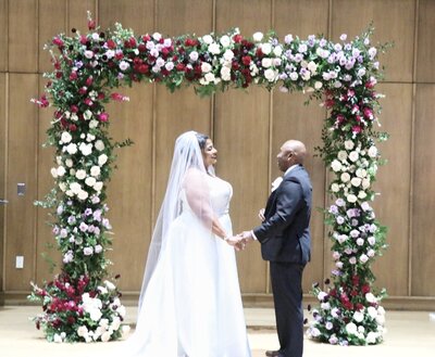 A heartfelt wedding ceremony moment, where a couple exchanges vows beneath a lush floral chuppah filled with soft pink, white, and deep red roses, creating a romantic ambiance.