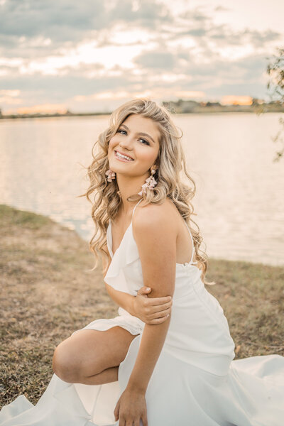 senior girl wearing a long white dress smiling at the camera and sitting near a pond at sunset