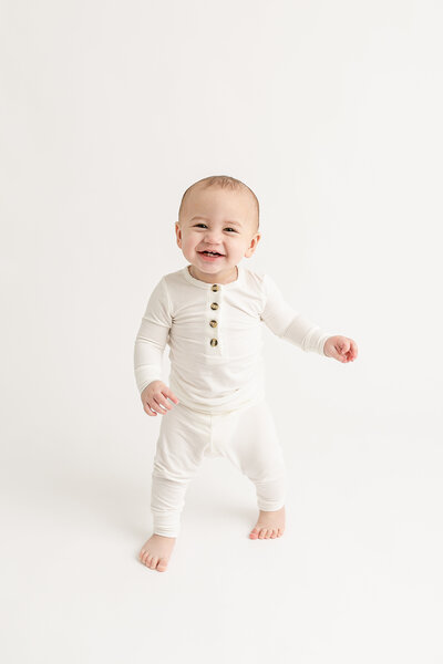 10 month old baby standing and smiling at the camera wearing an all white top and bottoms from Lou Lou and Company. In a portland oregon all white portrait studio.