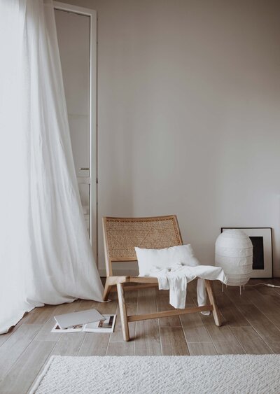 white walls, flowing white drapes, wood and rattan chair with white pillow, picture frame on floor, white vase on floor, wood floors