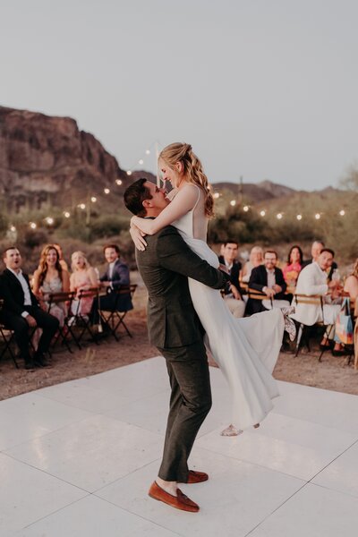 Groom lifts bride into the air during their first dance in the desert at sunset