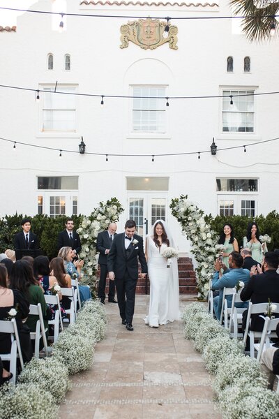 A joyful bride and groom walk hand-in-hand down the aisle moments after their wedding ceremony at the historic Casa Marina Hotel in Jacksonville, Florida. The outdoor courtyard is beautifully styled with lush white floral arrangements and baby's breath lining both sides of the aisle, creating a romantic and elegant atmosphere. Guests seated on white chairs cheer and applaud as the couple passes by, beaming with happiness. Twinkling string lights hang above, adding charm to the soft, coastal ambiance. This timeless moment was captured by a Jacksonville wedding photographer, known for documenting elegant and true-to-color celebrations.