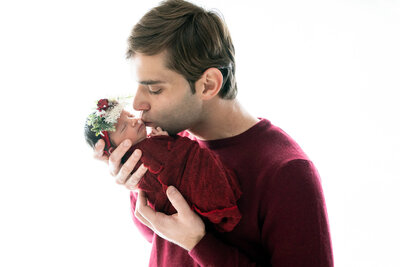 Dad holding newborn girl in front of a white background, both are wearing deep red and he is leaning down to kiss her