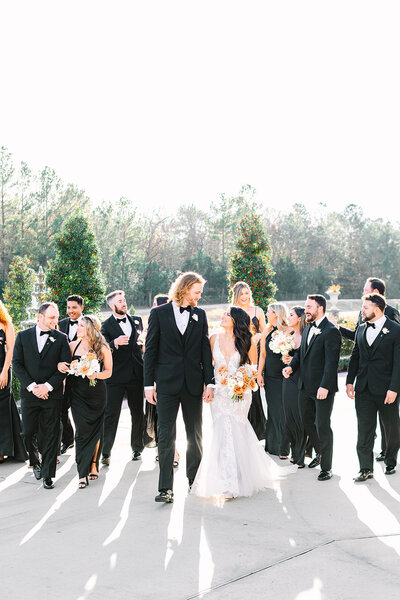Bride and groom holding hands smiling at each other with bridal party in the background
