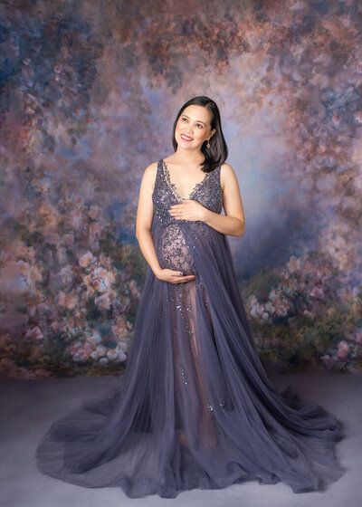 Maternity photography -- Pregnant woman in a beaded lavender gown poses against a painterly floral backdrop in a Birmingham maternity photography studio