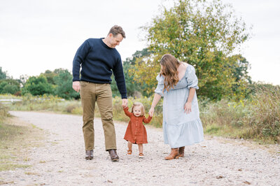 Mom and dad hold baby girls hands while walking on gravel path during family photos by Worth Capturing