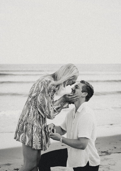 Groom-to-be proposing on the beach as his partner smiles and holds his face during their Portland Maine engagement.