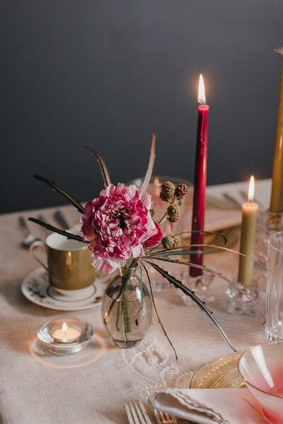 Tall red taper candle with small vase of pink flowers on a table with china cups and saucers designed by Jessamine Floral & Events