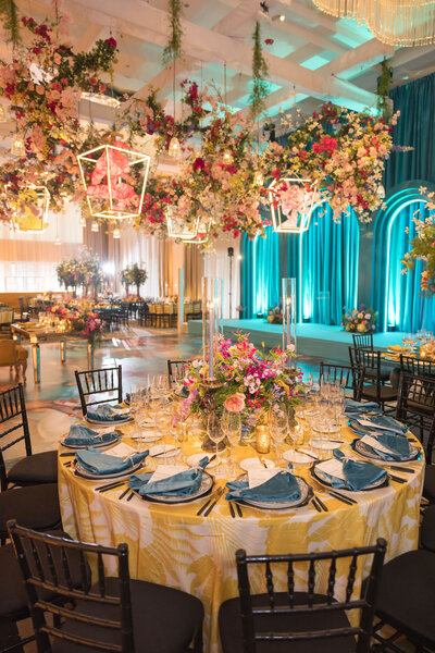 Blue yellow pink and white colorful wedding reception with round tables, yellow floral linen, blue napkins and uplighting along the room