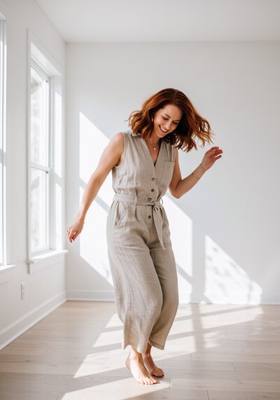 Woman joyfully dances barefoot in a sunlit room, wearing a beige jumpsuit. Bright windows and wooden floor create a serene and happy atmosphere.