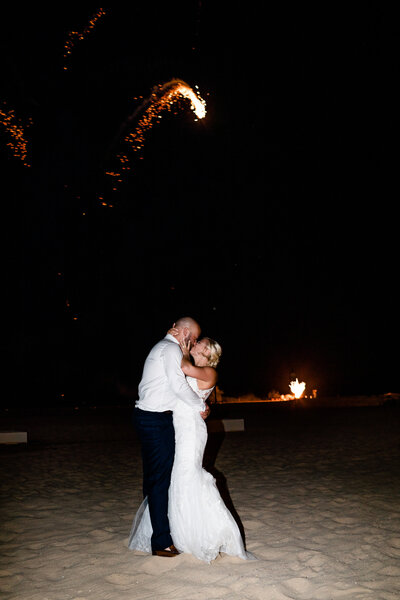 A bride and groom share a kiss on the beach at night while fireworks light up the sky above them. The romantic nighttime scene captures the intimacy and celebration of a destination wedding.
