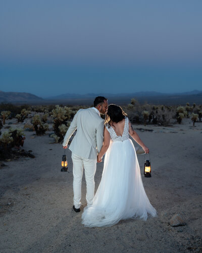 Bride and groom eloping at Joshua Tree National Park at Cholla Cactus Garden during vibrant colorful blue hour holding lanterns and kissing.