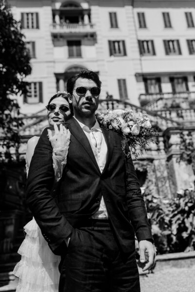 Black and white editorial-style portrait of a bride and groom in sunglasses, posing confidently in front of a European estate in Lake Como, Italy