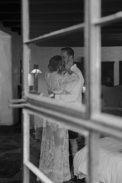 Bride and groom seen through a windowpane inside a cozy room.
