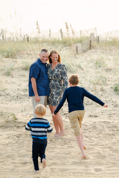 A couple puts their arms around each other on the beach at the Outer Banks. Their two sons run towards them. The family is dressed in blues and khakis.