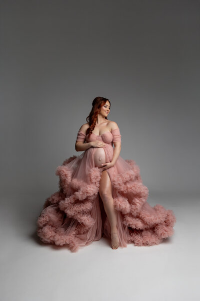 Pregnant woman in a flowing blush pink tulle gown standing barefoot in a studio, cradling her baby bump with a serene expression during her maternity session. Photograph by Yucaipa maternity photographer Kaitlyn Dawn Photography.