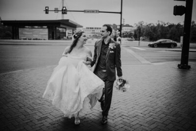 A joyful bride and groom walk hand-in-hand across a street near Bridge Park in Dublin, Ohio, their happiness captured in a timeless black and white photo. The bride, in a classic off-the-shoulder wedding gown, looks adoringly at the groom who is smartly dressed in a tailored gray suit with a flower boutonniere, embodying the elegance and excitement of their Dublin, Ohio wedding day.
