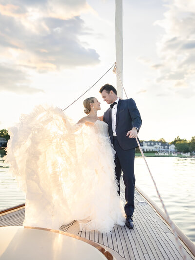 A couple stands on a boat at sunset. The woman is in a voluminous white wedding gown, and the man is in a black tuxedo. They gaze at each other lovingly, with the serene water and a house in the background.