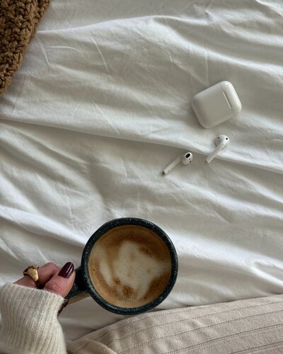 female virtual assistant in a lounge set sitting on her bed holding a coffee mug next to airpods
