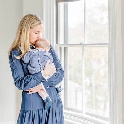 A mom in a blue dress holds her baby and looks out the window in Boston