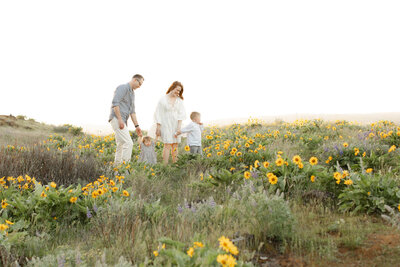 Dad, toddler girl, Mom and 4 year old boy are all dressed in light tones of white, grey and light blue and are walking hand in hand in single file through a field of short yellow wildflowers at Rowena Crest in Mosier Oregon in the Columbia River Gorge. They are all smiling as the little boy leads them through the flowers