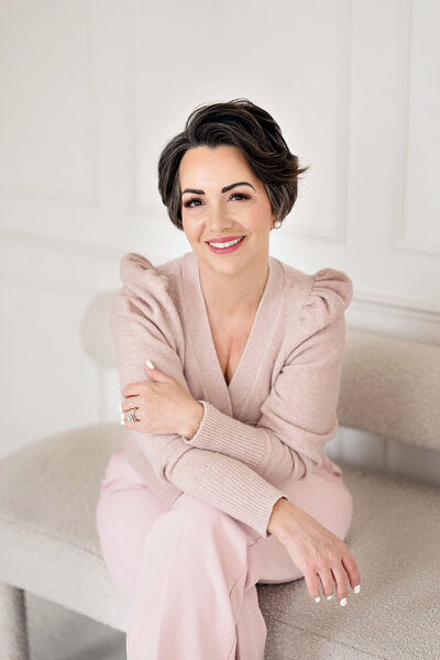 Kari Dawson, lead photographer of Kari Dawson + Co, seated with arms gently crossed in a soft pink sweater and trousers, smiling with warmth and elegance, reflecting on the quiet moments that matter most