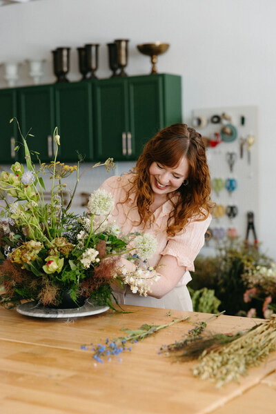 Mary Love standing in from of fall reception table floral arrangements in colors of mauve, cream, dusty rose, dusty blue, taupe, and natural green. Autumn floral consisting of roses, rain tree pods, fall branches, clematis, and greenery. North Carolina wedding design by Rosemary and Finch floral design based in Nashville, TN.
