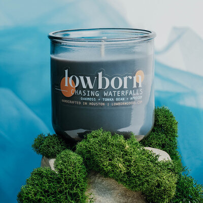 The chasing waterfalls candle, propped on a pile of rocks covered in moss. The background is an airy blue tulle. The image evokes calming waves, mossy riverbanks, and briny seas.