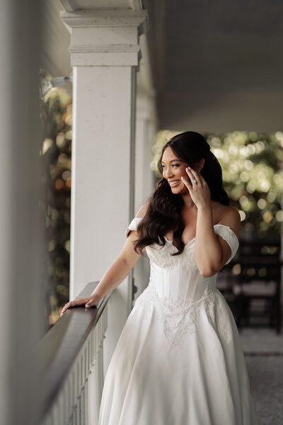 Bride looking off a balcony while pushing some hair behind her ear smiling at what she is looking at.