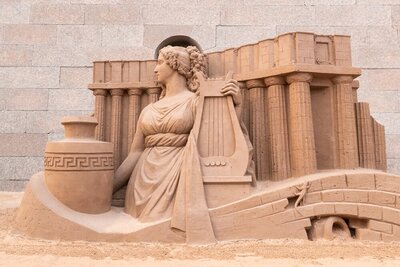 Temporary artwork made of sand depicts one of the Greek muses, holding a harp and standing in front of columns.