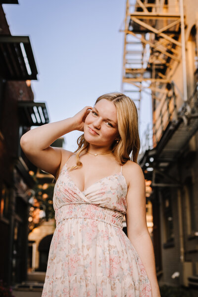 High school senior  in a floral dress standing in an alleyway with fire escapes.