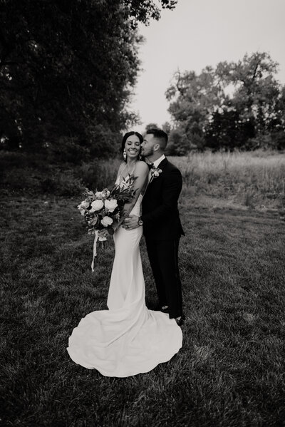 black and white image of bride backing into the groom for a kiss on the cheek while the bride holds her large timeless bouquet of flowers