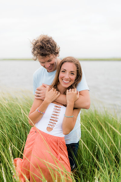 Best high school senior photographer in New Jersey.Picture of a couple hugging in the middle of a marsh field. The girl is wearing a long orange skirt and the man is wearing a baby blue tshirt. They are laughing.