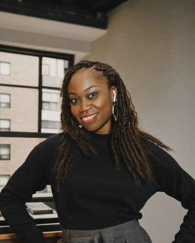 A professional portrait of a confident Black woman with shoulder-length twist braids and a warm smile. She is wearing a black turtleneck sweater and gray high-waisted pants. The background features a modern office environment with large windows showing an urban view.