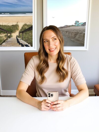 Stacey from Xanthe Bookkeeping smiling while sitting at a white desk holding an iPhone, with Outer Banks landscape photos on the wall behind her, used on the Xanthe Bookkeeping Contact page contact form section.