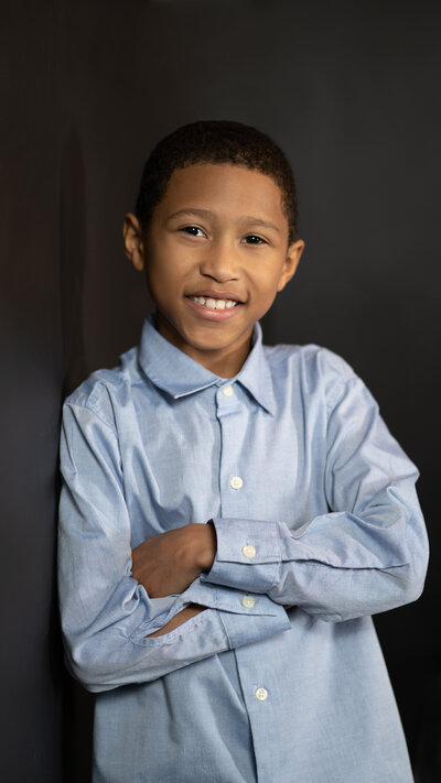 young boy smiles, dressed in blue shirts folds his arms and rests against a wall