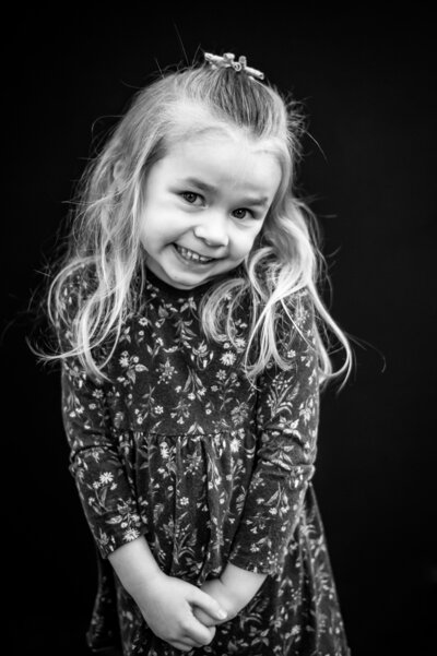 Little blonde girl in floral dress reluctantly smiling with right shoulder shrugged to chin in black and white | Meg Sivakumar Photography in Woodinville, WA