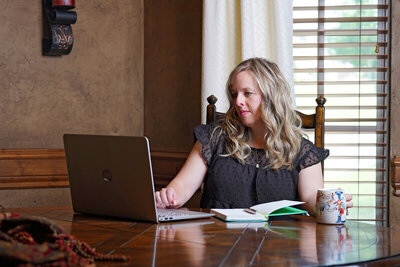 A woman sits at a wooden table, working on a laptop, possibly refining a Showit website for her personal brand. A notebook and a mug are in front of her, and she is wearing a black top. There is a window with blinds and curtains in the background.