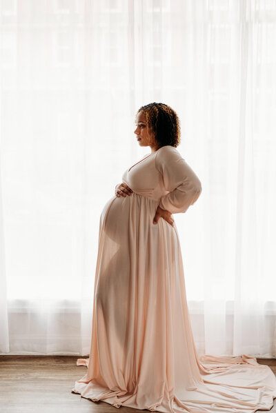 pregnant woman standing in front of a large window with thin drapes.  She is wearing a light pink dress, through which, you can see her silohuette.  She has one hand on her back hip, and the other resting on top of her baby bump.  Photo taken during a Delaware Maternity Photography session