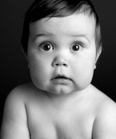A black and white photo of a baby with large, expressive eyes gazing curiously at the camera.