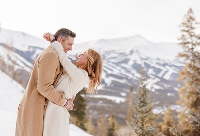 A happy couple in brown and tan winter coats hug and smile on a Breckenridge mountaintop