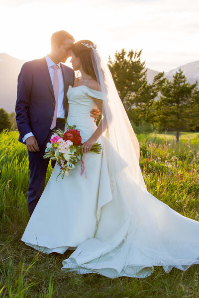 A bride in an off-the-shoulder white gown with a long train stands with a groom in a navy suit at Jackson Hole Golf and Tennis Club. She holds a bright bouquet of red, pink, and white flowers, while her hairpiece and flowing veil catch the evening sunlight. They lean in close, framed by vibrant green grass and the majestic Grand Tetons in the background.