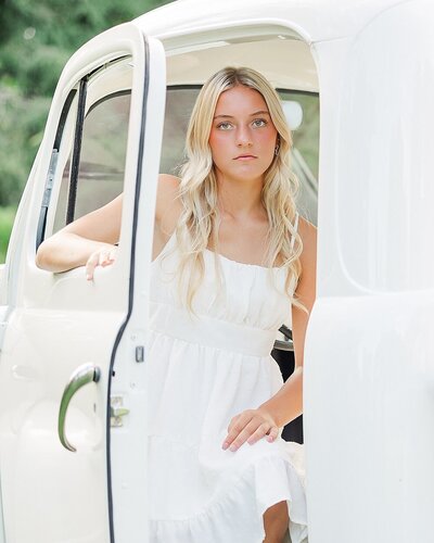 Senior girl with a white dress sitting in the front of a truck at a North East, PA, outdoor senior portrait location.