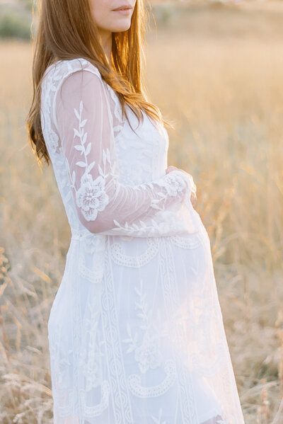Pregnant woman stands in golden grass field wearing long sleeved lace dress with golden sun shining through her hair and outlining her pregnant belly by Portland Newborn Photographer Emilie Phillipson Photography