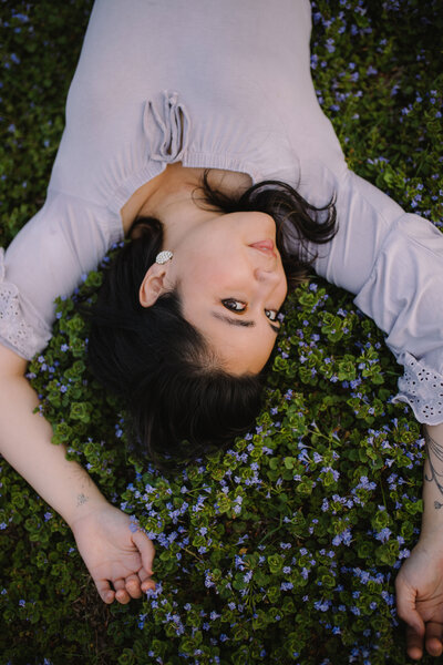 A woman in a light blouse lies on her back in a field of small purple flowers, arms outstretched and head turned gently to the side, gazing upward with a serene expression.