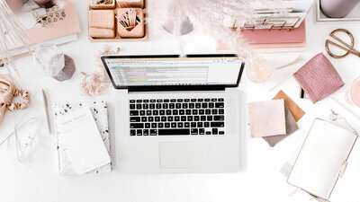 Flatlay of a laptop, planner, and desk accessories styled in soft pink and neutral tones, representing done-for-you HoneyBook workflows for wedding photographers.