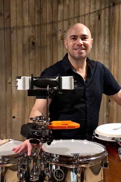 Percussionist smiling behind a timbales drum kit with a wooden backdrop, ready for a live wedding performance.