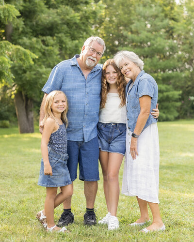 Grandparents and granddaughters standing next to each other with their arms around each other smiling at the camera, standing in grass with trees in the background