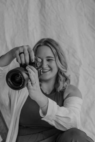 Smiling woman with glasses leans on a wooden chair. The background has framed art including a butterfly. The mood is warm and nostalgic.