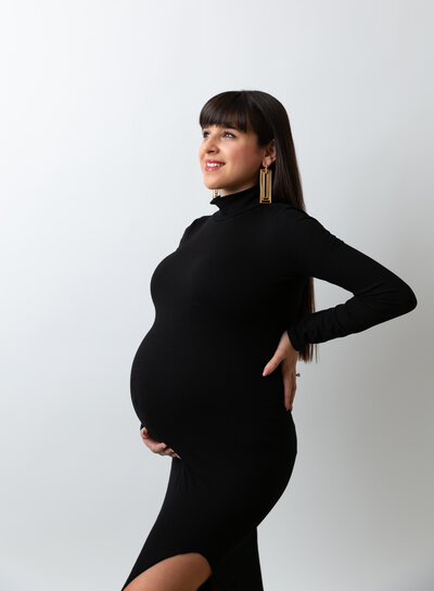 Expectant mom in fitted black grown poses for maternity photos in Brooklyn, NY photo studio. She has one hand on her back and one hand under her bump. She is looking up at the light and smiling.
